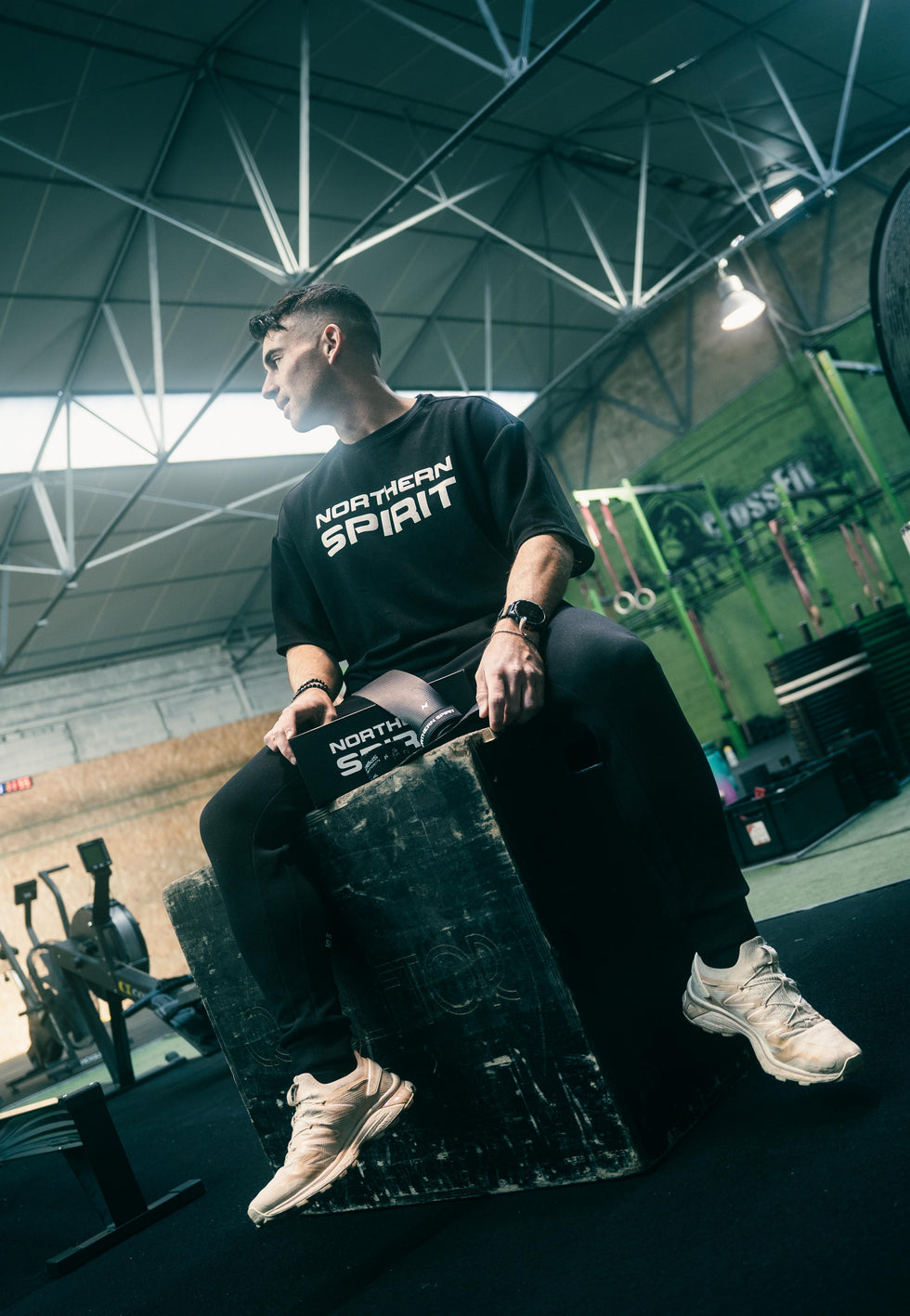 Athlete wearing a black Northern Spirit oversized T-shirt and training pants, sitting on a plyo box in a CrossFit gym — premium functional training apparel.