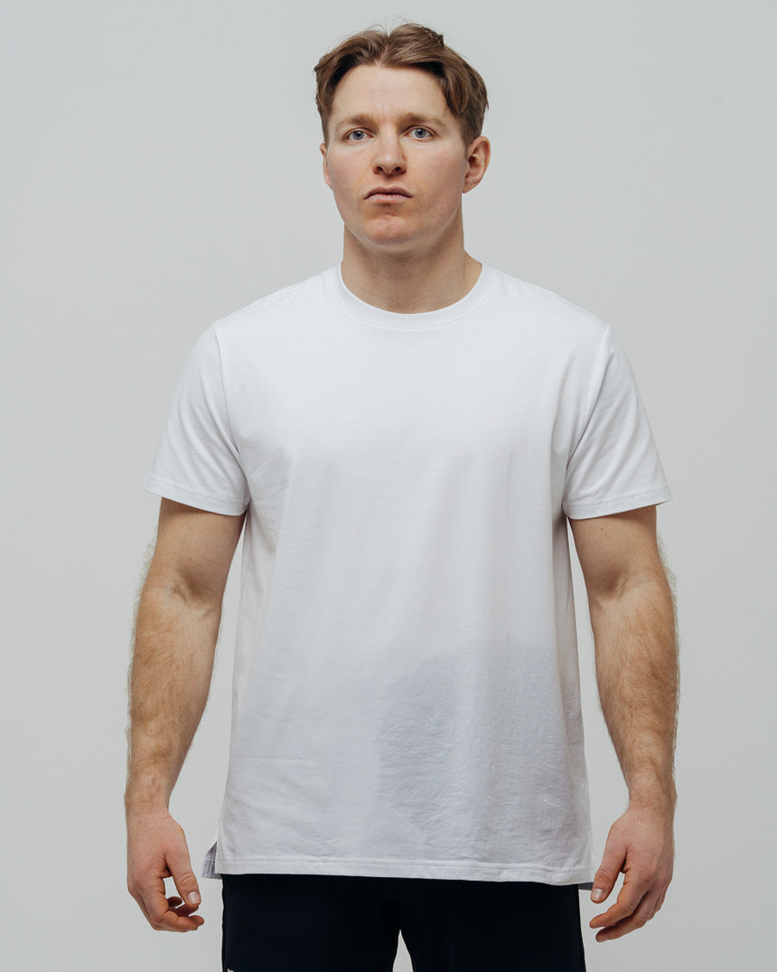 A man wearing a plain white T-shirt and black pants standing against a neutral background.
