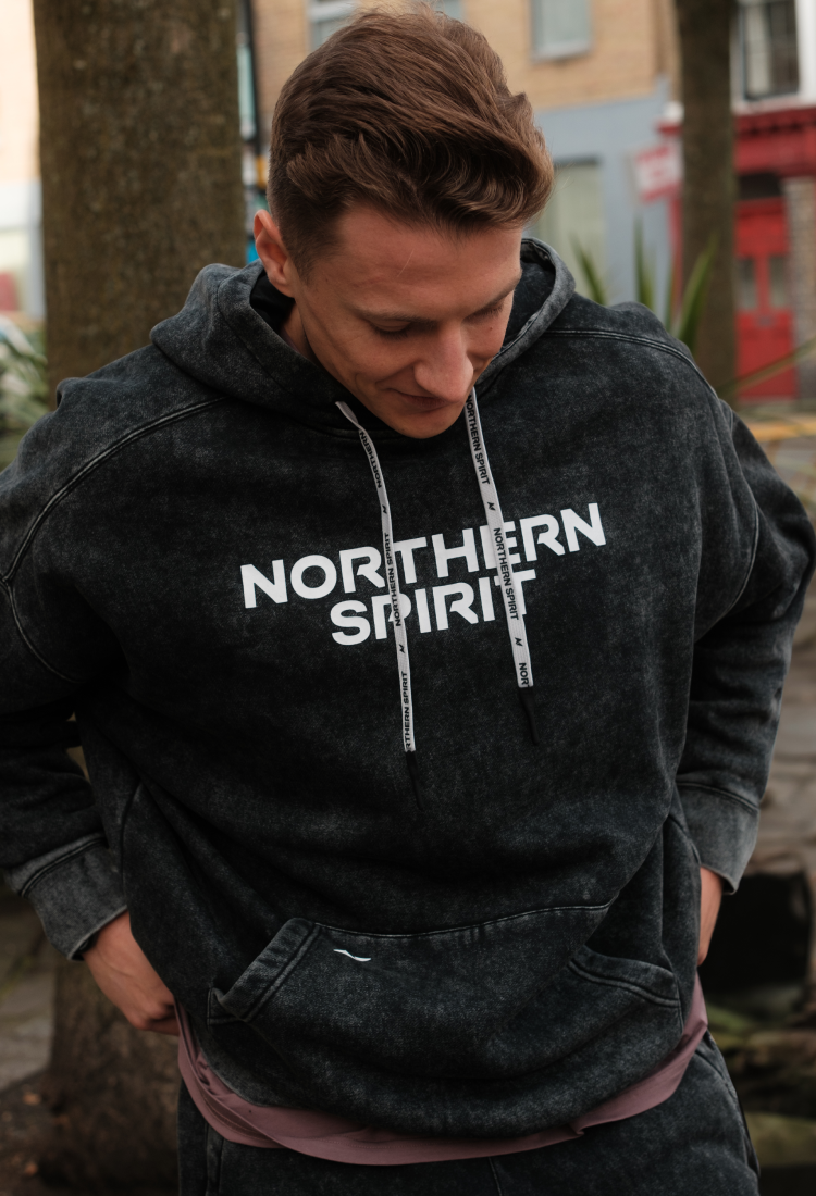 Male athlete wearing a black washed Northern Spirit hoodie looking down with hands in pockets – Cozy Collection – premium CrossFit and lifestyle hoodie for comfort and recovery.