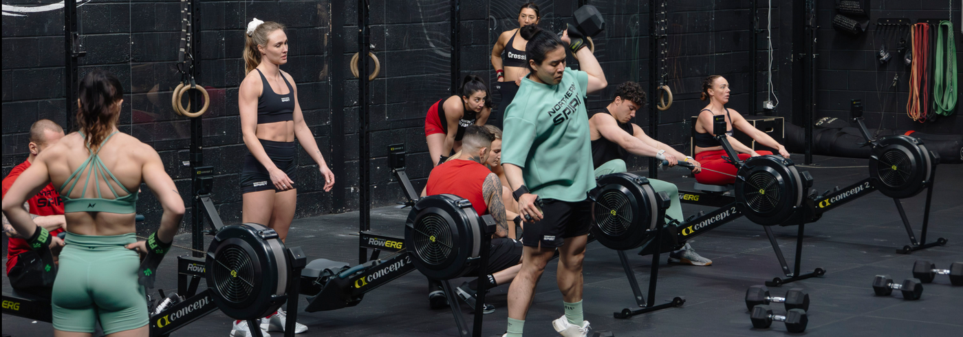 Group of CrossFit® athletes training in a functional fitness gym wearing Northern Spirit apparel during a team workout with Concept2 rowers and dumbbells.