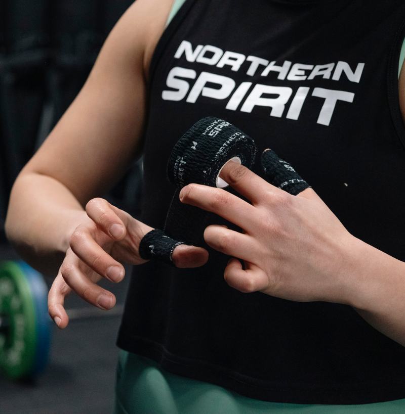 Female CrossFit® athlete applying Northern Spirit black grip tape on fingers while wearing black tank top and green leggings in a gym.
