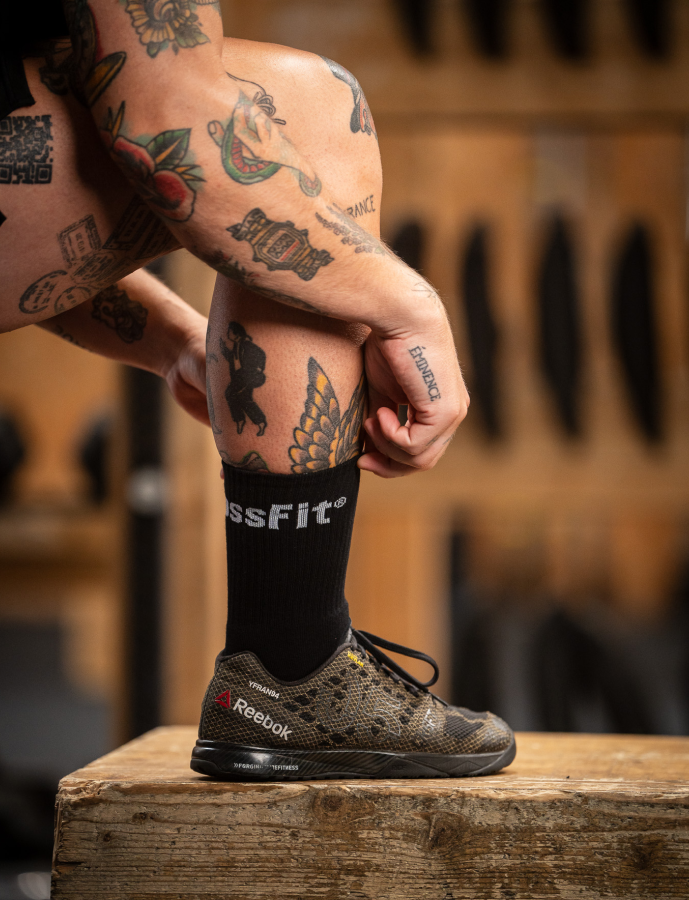 Person wearing Reebok shoes and CrossFit socks with tattoos on arms and legs, standing on a wooden block.